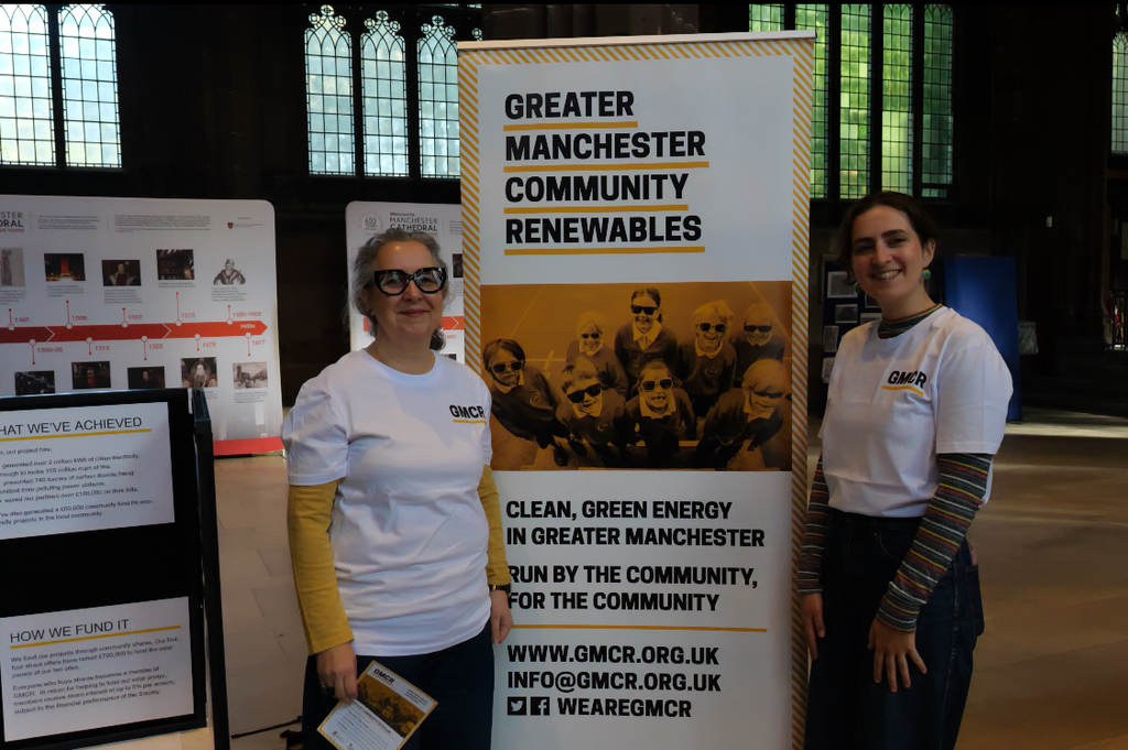 Photograph of GMCR volunteers Sarra and Louisa stood in front of a banner promoting GMCR